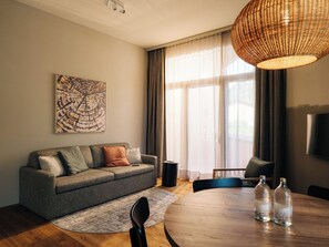 Ein modernes Wohnzimmer mit einem grauen Sofa, einem runden Holztisch mit Flaschen, einer gewobenen Leuchte und einem großen Fenster mit Vorhängen.