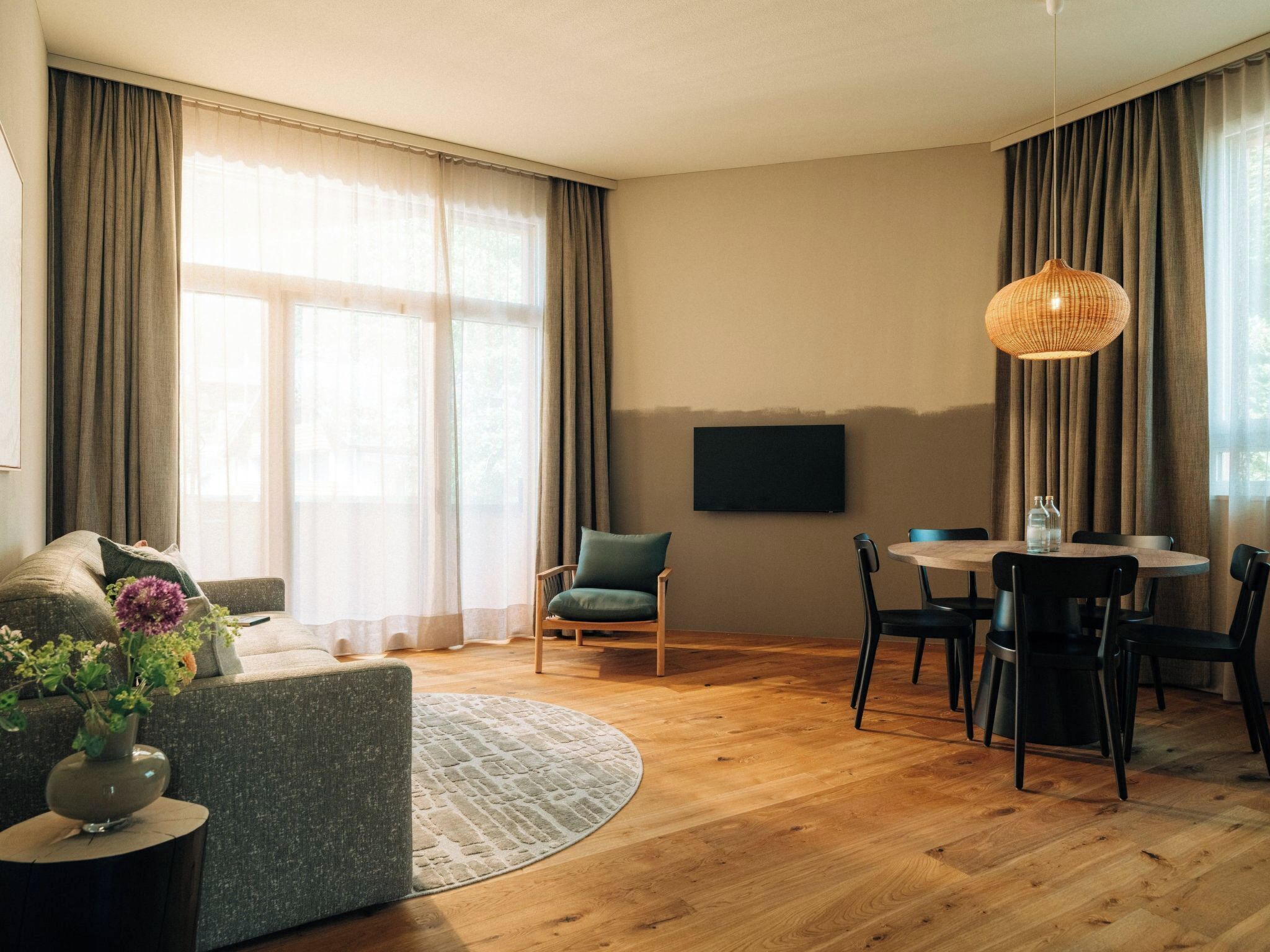 Ein modernes Wohnzimmer mit einer Couch, einem Esstisch mit Stühlen, einem an der Wand montierten Fernseher und großen Fenstern mit Vorhängen.
