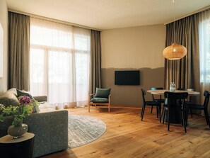 A modern living room with a sofa, a dining table with chairs, a TV mounted on the wall, and large windows with curtains. - KURVE Apartments & Lounge Appartement/Fewo, Bad, WC, 2 Schlafräume by Interhome (Adelboden)