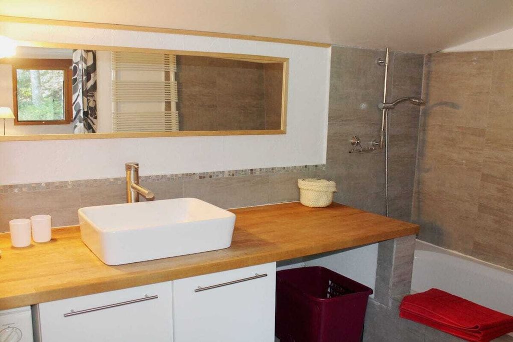 Ein Badezimmer mit einem weißen Waschbecken, einer Holzablage und einem Spiegel darüber. Es gibt einen Duschbereich mit Glastür und einem roten Handtuch auf dem Boden.
