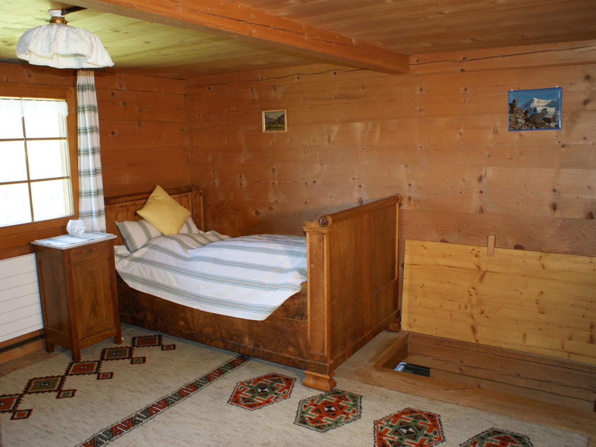 Quarto de madeira com cama, mesa de cabeceira, abajur e janela.