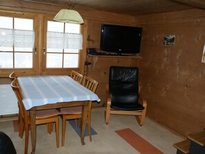 A room with a wooden interior, a table with chairs, a television mounted on the wall, and a window with a curtain.