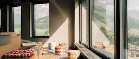 A modern interior with large windows, wooden furniture, and a view of a mountainous landscape.