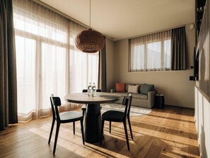 Un moderno salón con una mesa de comedor redonda, dos sillas, un sofá y un amplio ventanal con cortinas translúcidas.