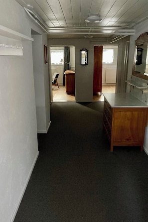 Ein Zimmer mit Teppichboden, einem Holzschrank, einem Schreibtisch und einem Stuhl. Es gibt ein Fenster mit Vorhängen, eine Heizung und eine Uhr an der Wand.