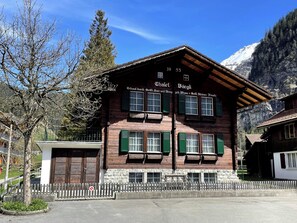A traditional chalet with a wooden facade, green shutters, and a sign indicating it is a 'Whalet' with a view of mountains in the background. - Chalet Bärgli Appartement/Fewo, Bad, WC Chalet Bärgli – Ferienwohnung Gemmi by Interhome (Kandersteg)