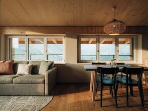 Ein gemütliches Wohnzimmer mit einer Couch, einem Esstisch mit Stühlen und großen Fenstern mit Blick auf die Berge.