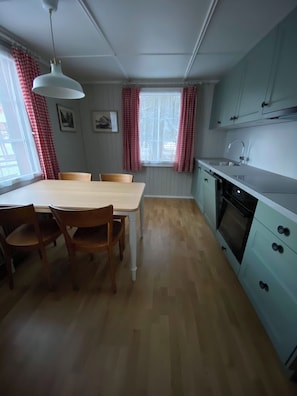 Eine kompakte Küche mit Esstisch, Stühlen, einer Pendelleuchte, einem Fenster mit Vorhängen und einem eingebauten Backofen.
