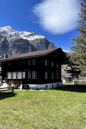 Ein traditionelles Chalet mit geneigtem Dach, einer Holzterrasse und Blick auf schneebedeckte Berge.