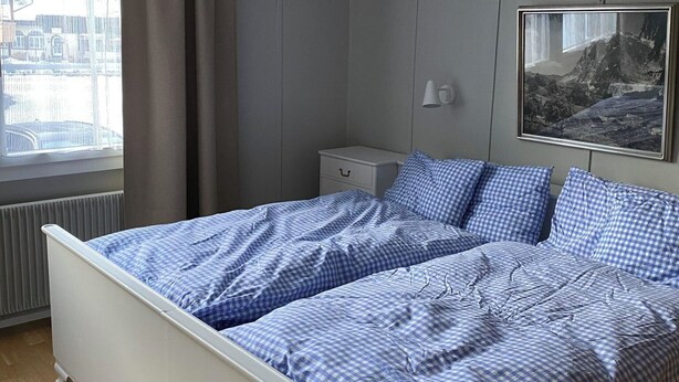 Un dormitori amb un llit, una tauleta de nit, una làmpada, una finestra amb cortines i un quadre al mur.