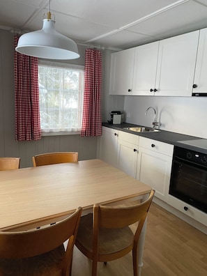 A kitchen with white cabinets, a black countertop, a sink, and an oven. There is a dining table with chairs and a window with red and white checkered curtains. - Chalet Bärgli Appartement/Fewo, Bad, WC Chalet Bärgli – Ferienwohnung Doldenhorn by Interhome (Kandersteg)