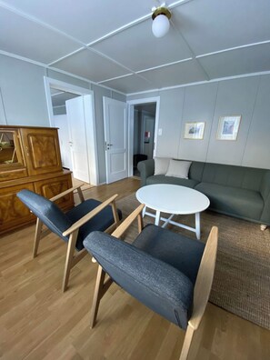 Ein Zimmer mit einem Holzkabinett, zwei Stühlen, einem runden Tisch, einer Couch und einer Deckenleuchte.
