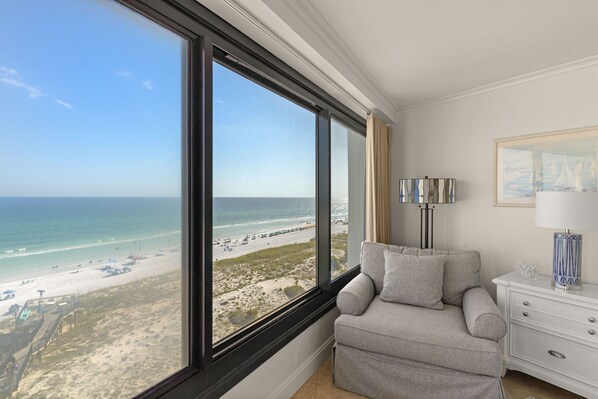 Een kamer met een groot raam met uitzicht op een strand, een grijze bank, een witte nachtkastje met een lamp, en een lijst met een foto aan de muur.