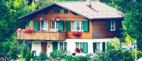 Une maison en bois à deux étages, dotée d’un balcon et entourée d’une végétation luxuriante.