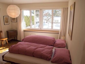 Ein Schlafzimmer mit Bett, Heizkörper, Fenster mit Vorhängen, Stuhl und Pendelleuchte.
