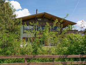 Ein mehrstöckiges Holzhaus mit geneigtem Dach, umgeben von üppiger Vegetation und einem Holzzaun.