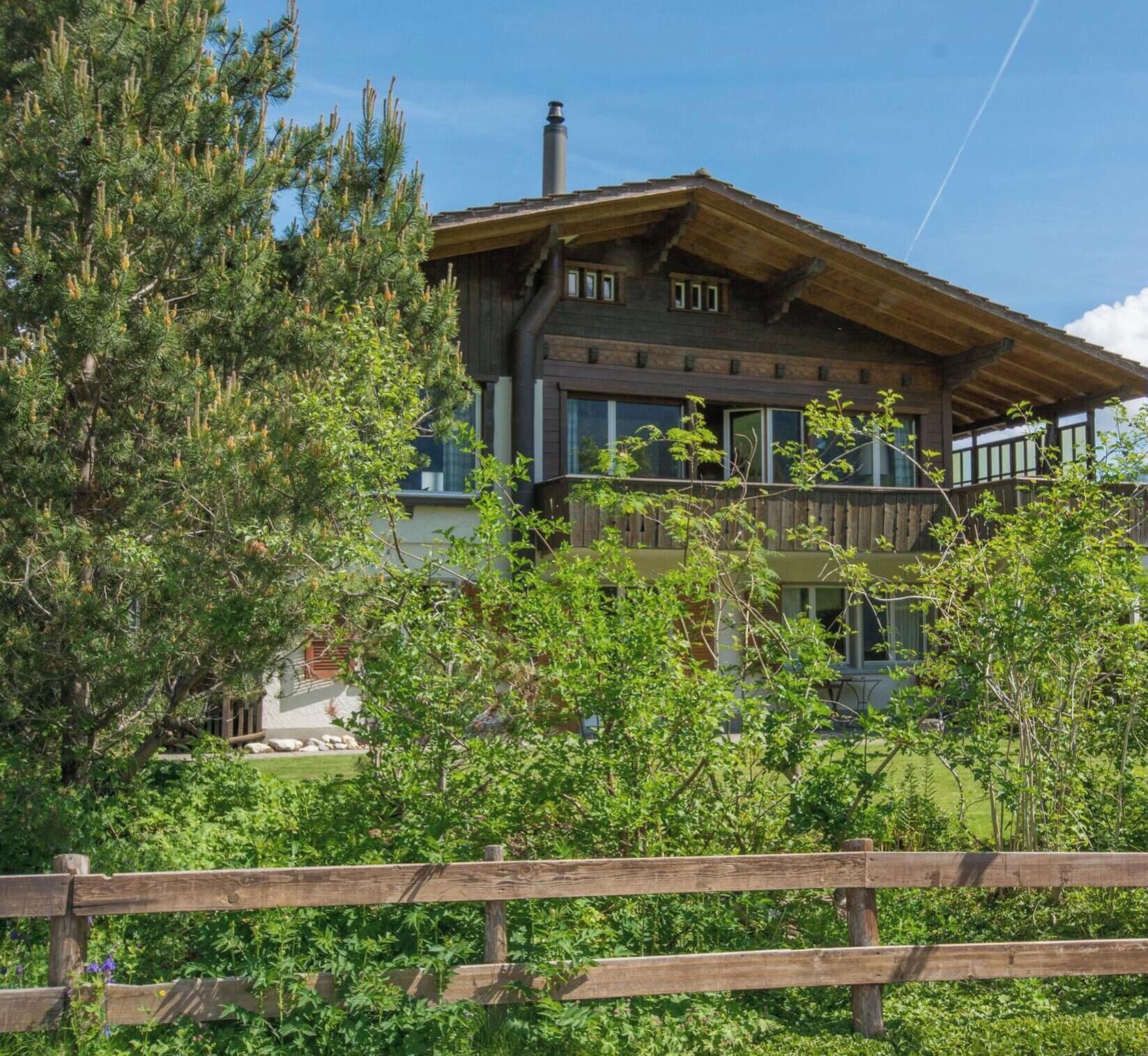 A two-story wooden house with a balcony, surrounded by greenery and trees.