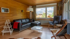Un saló modern amb sostre de fusta, un sofà de pell negra, una taula de cafè blanca, un butac armchair gris i vistes a les muntanyes a través de grans finestres.
