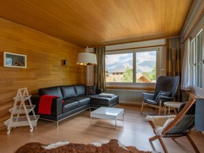 A modern living room with a wooden ceiling, a black leather sofa, a white coffee table, a grey armchair, and a view of mountains through large windows. - Baldrian C14 Appartement/Fewo, Bad, WC, 3 Schlafräume 4.5- Zimmerwohnung by Interhome (Adelboden)