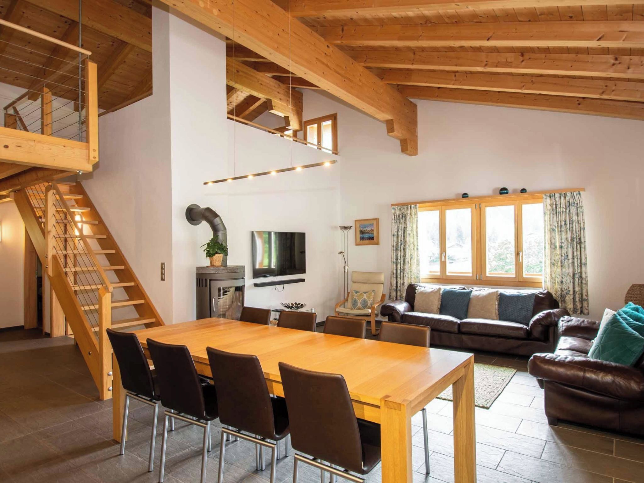 A spacious living area with a wooden staircase, a dining table with chairs, a sofa, and a television.