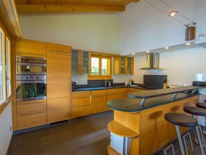 Una cuina moderna amb armaris de fusta, forn integrat, finestra gran i zona de bar amb tamborets.