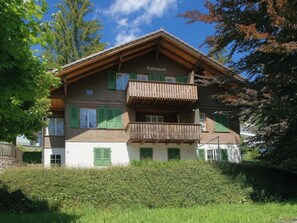 Ein zweigeschossiges Gebäude mit einem Balkon im oberen Stockwerk, umgeben von viel Grün.