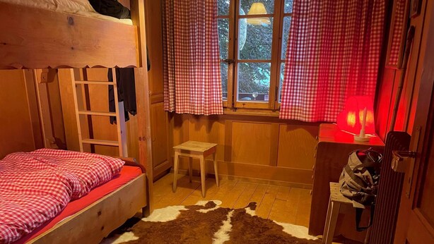 Ett mysigt rum med en våningssäng, ett litet träbord, en stol, ett fönster med gardiner och en röd lampa.