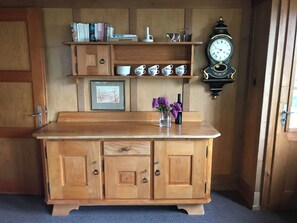 Eine Holzkommode mit mehreren Schubladen und Regalen, ein gerahmtes Bild, eine Vase mit Blumen und eine Wanduhr.