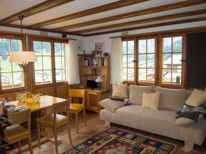 A cozy living room with a wooden ceiling, a sofa, a coffee table, a bookshelf, and large windows with curtains. - Chalet Zwirbeli Appartement/Fewo, Dusche, WC, 1 Schlafraum 2-Zimmerwohnung by Interhome (Adelboden)