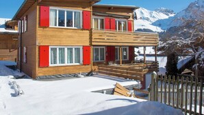 Eine zweistöckige Holzhütte mit roten Fensterläden, einem Balkon und Blick auf schneebedeckte Berge.