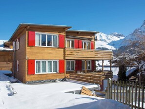 Eine zweistöckige Holzhütte mit roten Fensterläden, einem Balkon und Blick auf schneebedeckte Berge.
