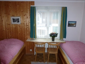 Ein Zimmer mit zwei Einzelbetten, einem Holztisch mit Stühlen, einem Fenster mit Vorhängen und zwei gerahmten Bildern an den Wänden.