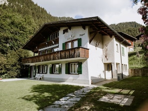 Ein zweistöckiges Gebäude mit einem Balkon, grünen Fensterläden und einem steinbelagten Weg zum Eingang.