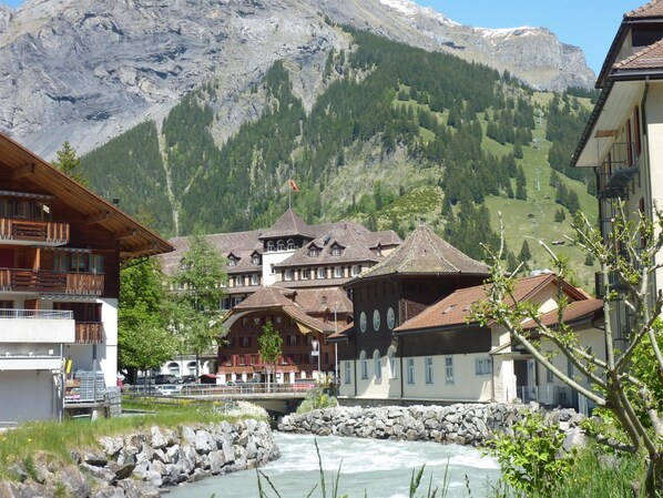 A riverside building with multiple balconies and a mountain backdrop.