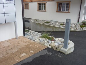 A building with a paved entrance, a stone retaining wall, and a small garden area with rocks and plants.