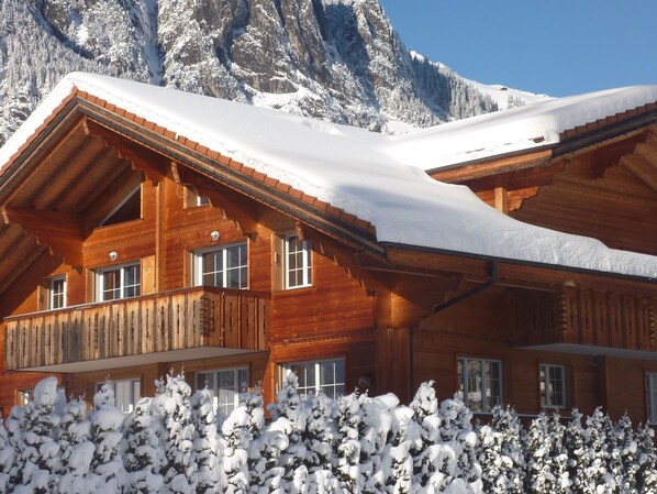 A wooden chalet with a sloped roof, surrounded by snow-covered trees and mountains in the background.