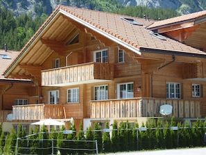 A multi-story wooden chalet with balconies, a sloped roof, and a mountainous backdrop.