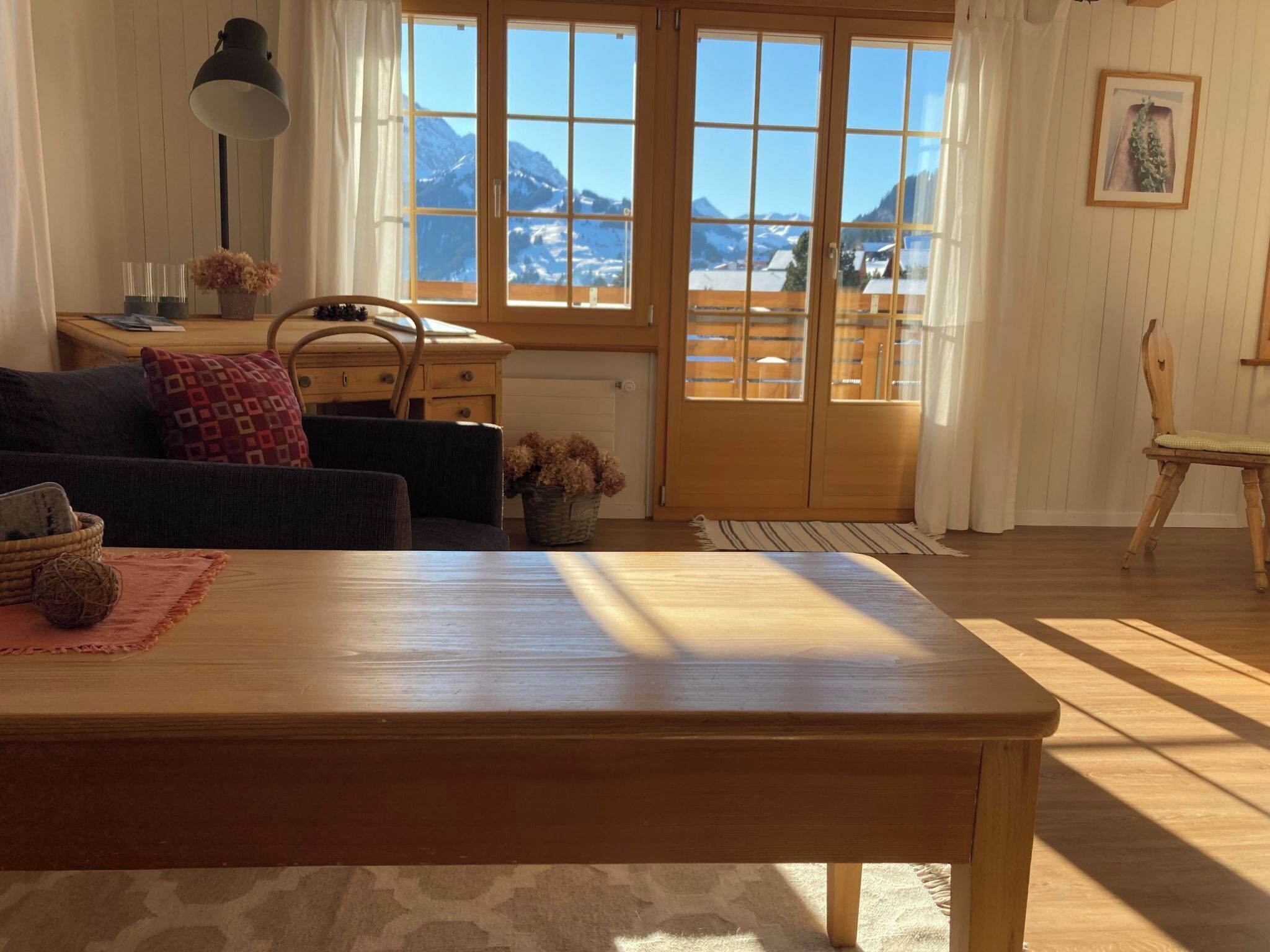 A cozy living room with a wooden table, a sofa, a desk, and a view of mountains through large windows.