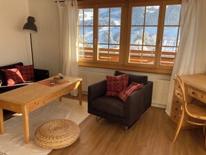 Ein gemütliches Zimmer mit einem Holztisch, einem dunklen Sessel und Blick auf eine verschneite Landschaft durch große Fenster.