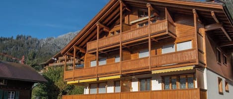 A multi-story wooden building with balconies, a sloped roof, and a mountainous backdrop.