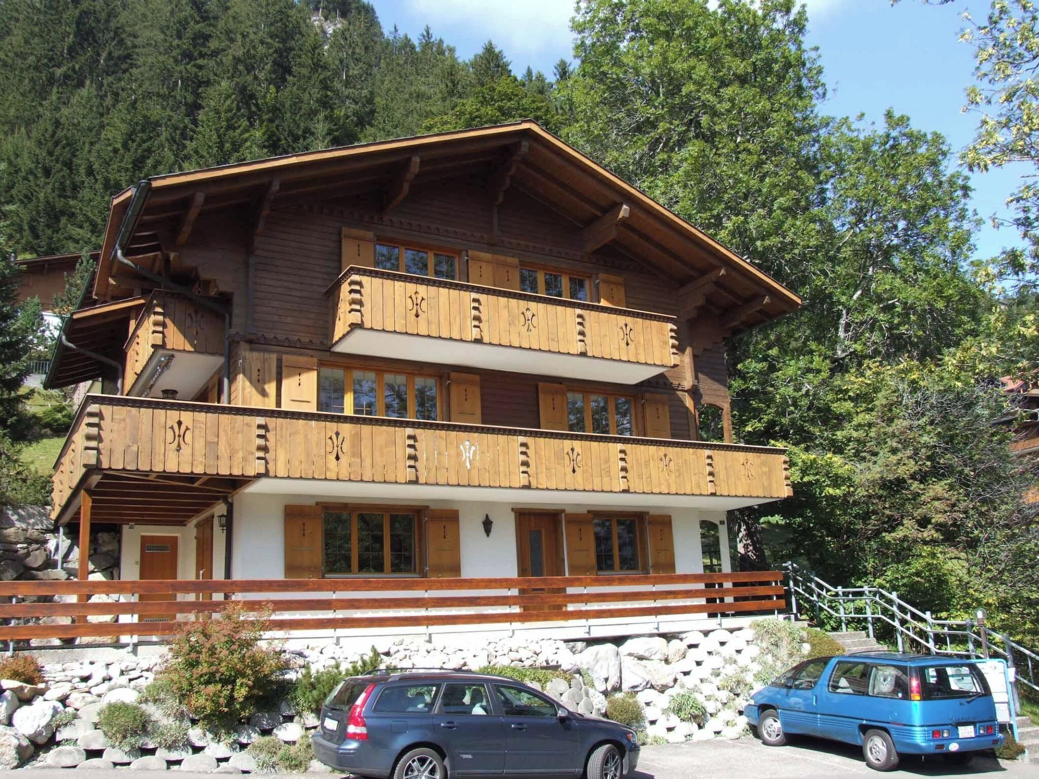 A multi-story wooden building with balconies, a stone staircase, and a parking area with cars.