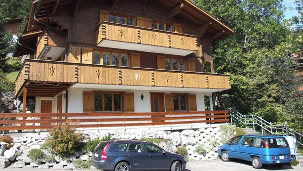 Ein mehrstöckiges Holzgebäude mit Balkonen, einer Steintreppe und einem Parkplatz mit Autos.