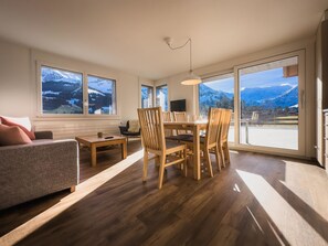 Ein geräumiges Wohnzimmer mit Holzboden, einer Couch, einem Esstisch mit Stühlen und großen Fenstern mit Blick auf die Berge.