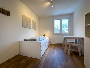 A compact bedroom with a bed, a desk, and a radiator. There are two windows and wall art. - Ferienwohnung Lohner (EG) Appartement/Fewo, Dusche, WC, 3 Schlafräume by Interhome (Adelboden)