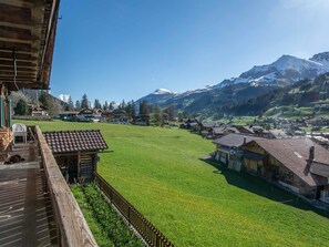 Ein Bergdorf mit traditionellen Holzhäusern, einer grünen Wiese und einem schneebedeckten Berg im Hintergrund.