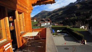 Een houten balkon met een tafel en stoelen, met uitzicht op een bergachtig landschap met huizen.