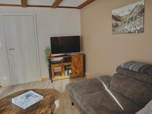 Una sala de estar con un sofá, una mesa de centro con revistas, un televisor sobre un mueble de madera y una pintura de montañas en la pared.