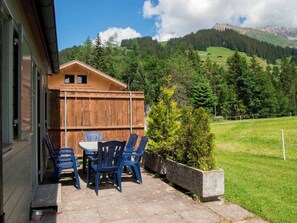 Une cabane en bois avec une table et des chaises sur une terrasse, entourée d’une pelouse et adossée à une montagne boisée.