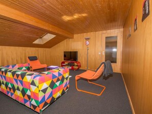 Ein Zimmer mit Holzboden, einem farbenfrohen, geometrisch gemusterten Sofa, einem kleinen orangefarbenen Sessel, einem Fernseher und einer Tür mit Spiegel.