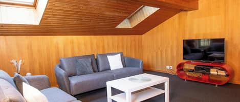 Un salon chaleureux avec un plafond en pente, un ensemble de canapés, une table basse et un téléviseur fixé à un mur en bois.
